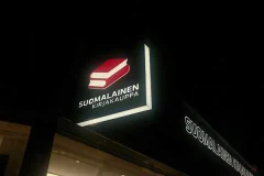 Suomalaisen Kirjakaupan valaistu kotelokyltti Suomalaisen Kirjakaupan punavalkoinen logokyltti valaistuna pimeää taivasta vasten