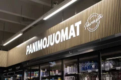 White illuminated Panimojuomat lettering on a wooden slat panel above drinks shelves White illuminated Panimojuomat lettering on a wooden slat panel above drinks shelves