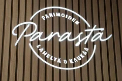 White circular illuminated lettering reading Panimoiden parasta läheltä ja kaukaa on a wooden slat wall
