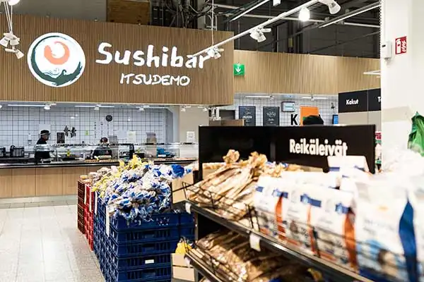 Sushibar Itsudemo sign on a wood-effect wall panel behind bread shelves in a grocery store