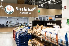Sushibar Itsudemo -kyltti puukuvioisessa seinäpaneelissa ruokakaupan leipähyllyjen takana