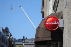 Veikkauksen pyöreä ulkokyltti Punainen pyöreä Veikkaus-kyltti ulkonee julkisivusta markiisin vieressä.