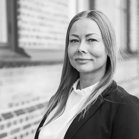 Svartvitt porträtt av Merja framför en tegelvägg och ett fönster
