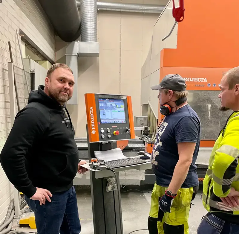 Kolme henkilöä tuotantokoneen oranssin ohjauspaneelin vieressä