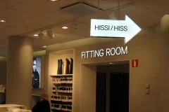 An illuminated Hissi/Hiss arrow and Fitting Room lettering above a store doorway. An illuminated Hissi/Hiss arrow and Fitting Room lettering above a store doorway.