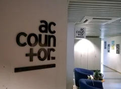 A black Accountor logo on a light office wall and a hanging sign further down the corridor. A black Accountor logo on a light office wall and a hanging sign further down the corridor.