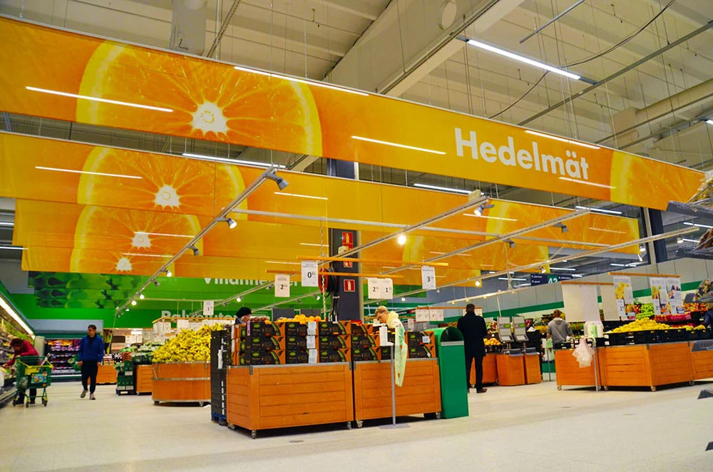 Oranssit hedelmäkuvat ja Hedelmät-teksti ruokakaupan hedelmäosaston yläpuolella.