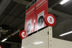 Red Tokmanni promotional signs and an aisle marker numbered 3 above a shelf. Red Tokmanni promotional signs and an aisle marker numbered 3 above a shelf.