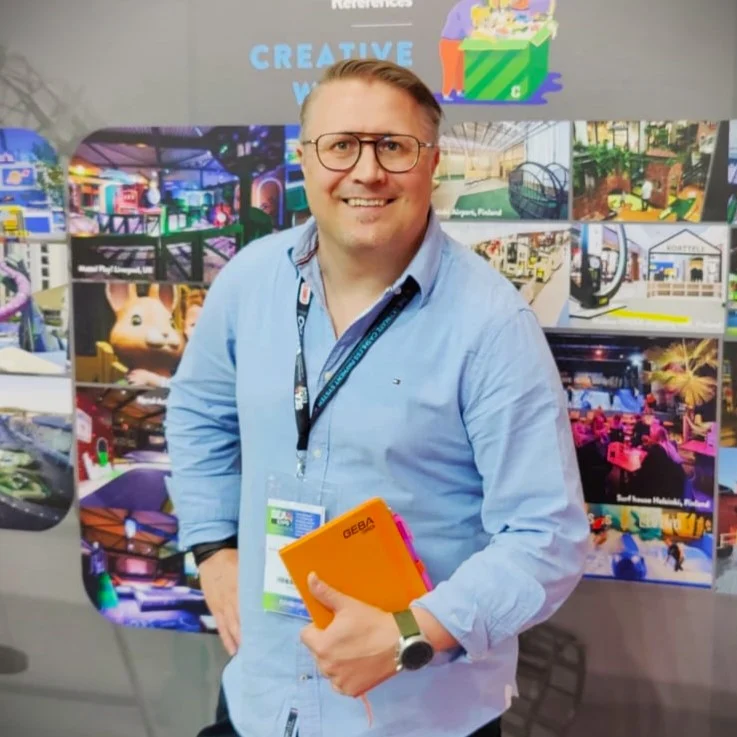 Jari Partanen vaaleansinisessä paidassa oranssi muistikirja kädessään kuvaseinän edessä