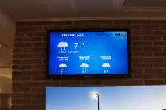 Sininen Kajaanin sääennuste tiiliseinään kiinnitetyllä näytöllä.