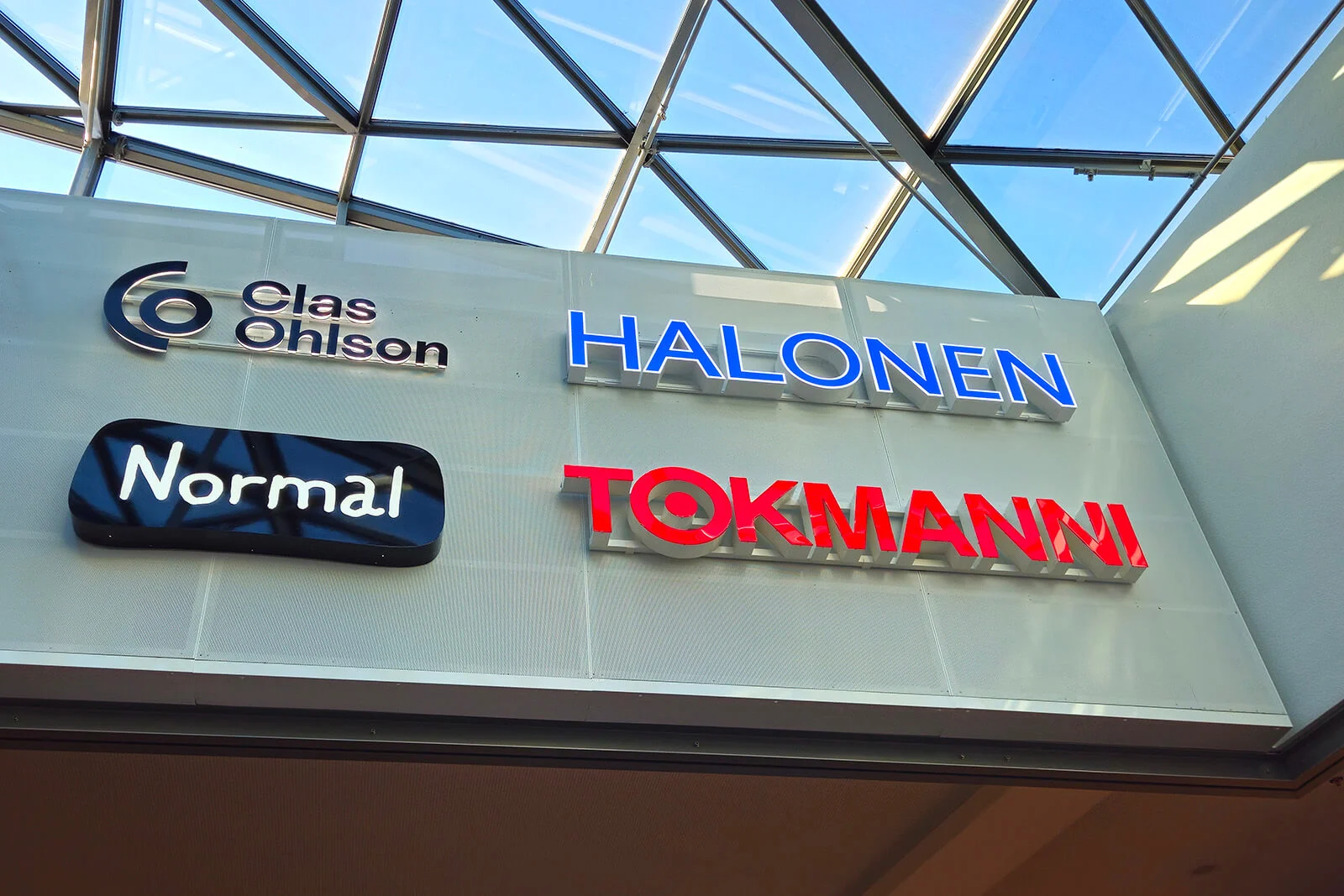 Clas Ohlsonin, Halosen, Normalin ja Tokmannin kyltit kauppakeskuksen seinällä lasikaton alla