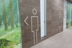 Large restroom pictograms and a left arrow on a wood-patterned wall.
