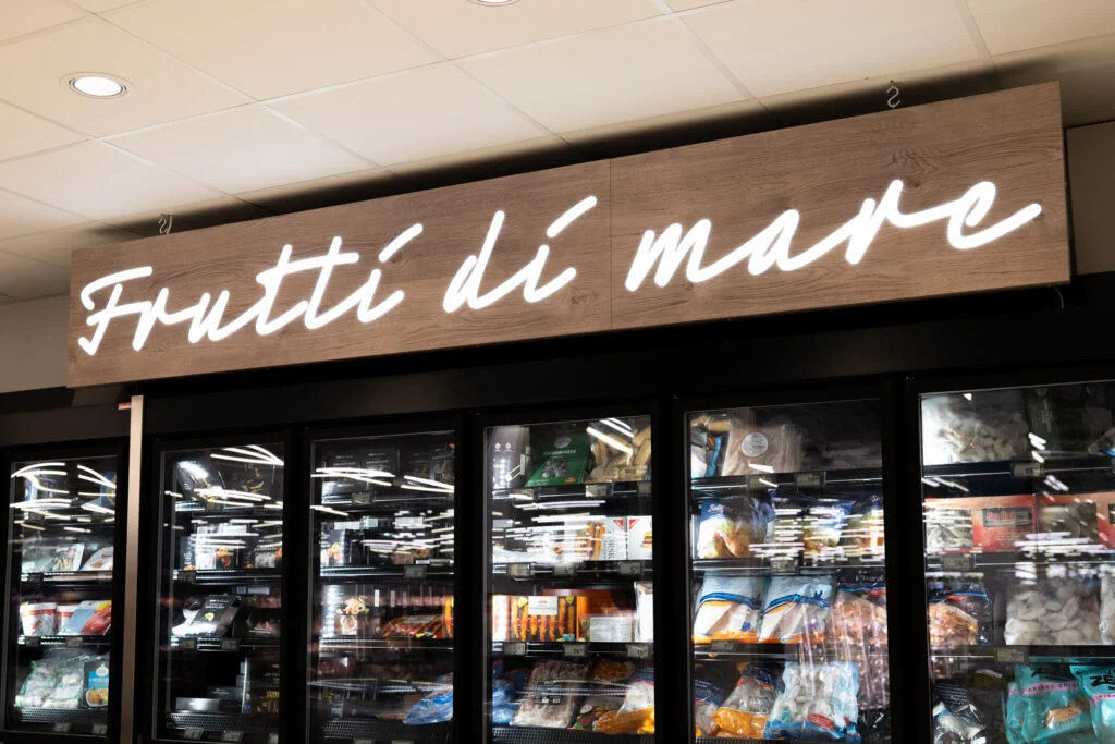 Valkoinen Frutti di mare -valoteksti puukuvioisessa opasteessa pakastekaappien yläpuolella Prisma Sellossa