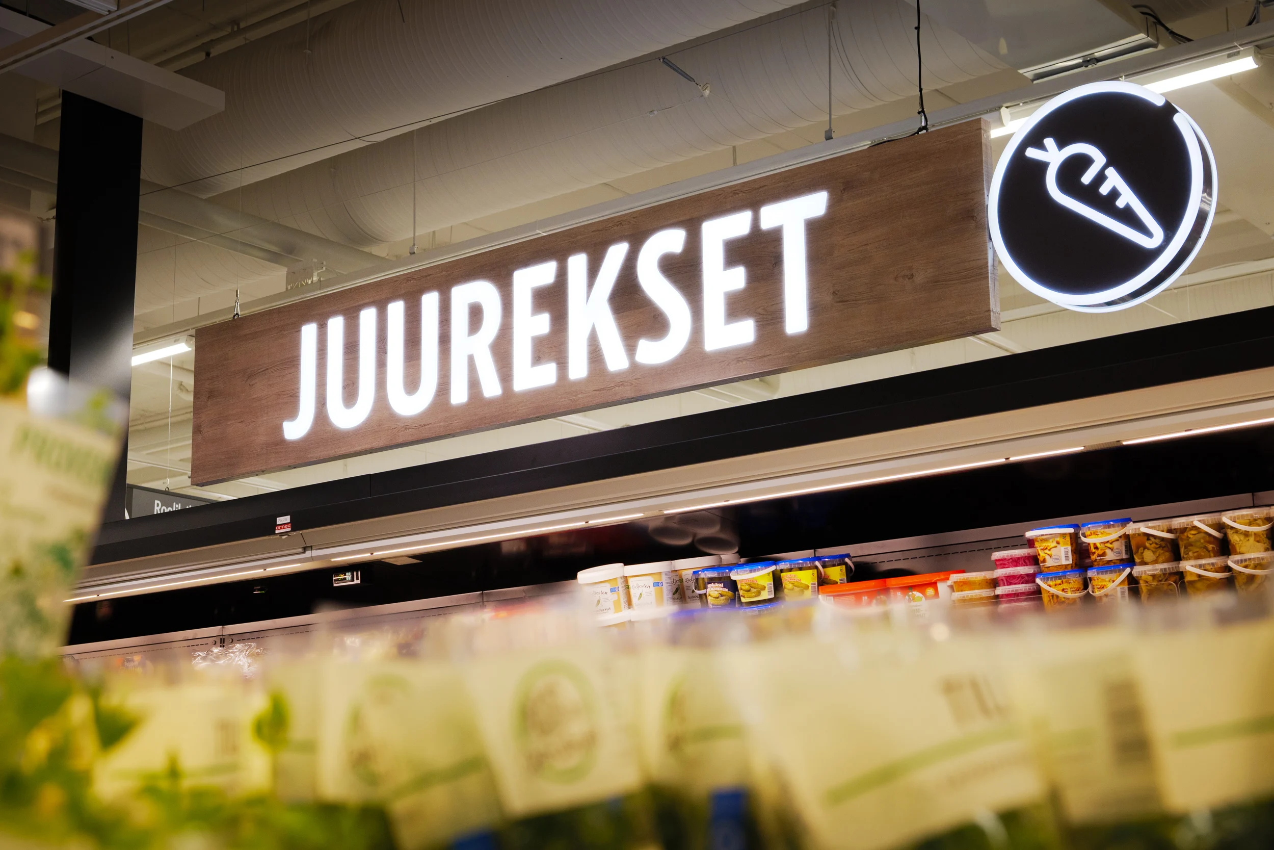 Valaistu Juurekset-teksti ja porkkanasymboli vihanneshyllyn yläpuolella Prisma Sellossa