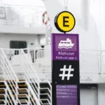 Violetti Rådhuset-opastepylväs, keltainen E-tunnus ja lautta taustalla.