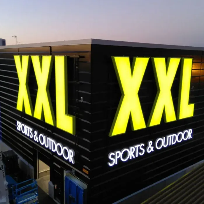 Large yellow illuminated XXL letters on two sides of a building at dusk.