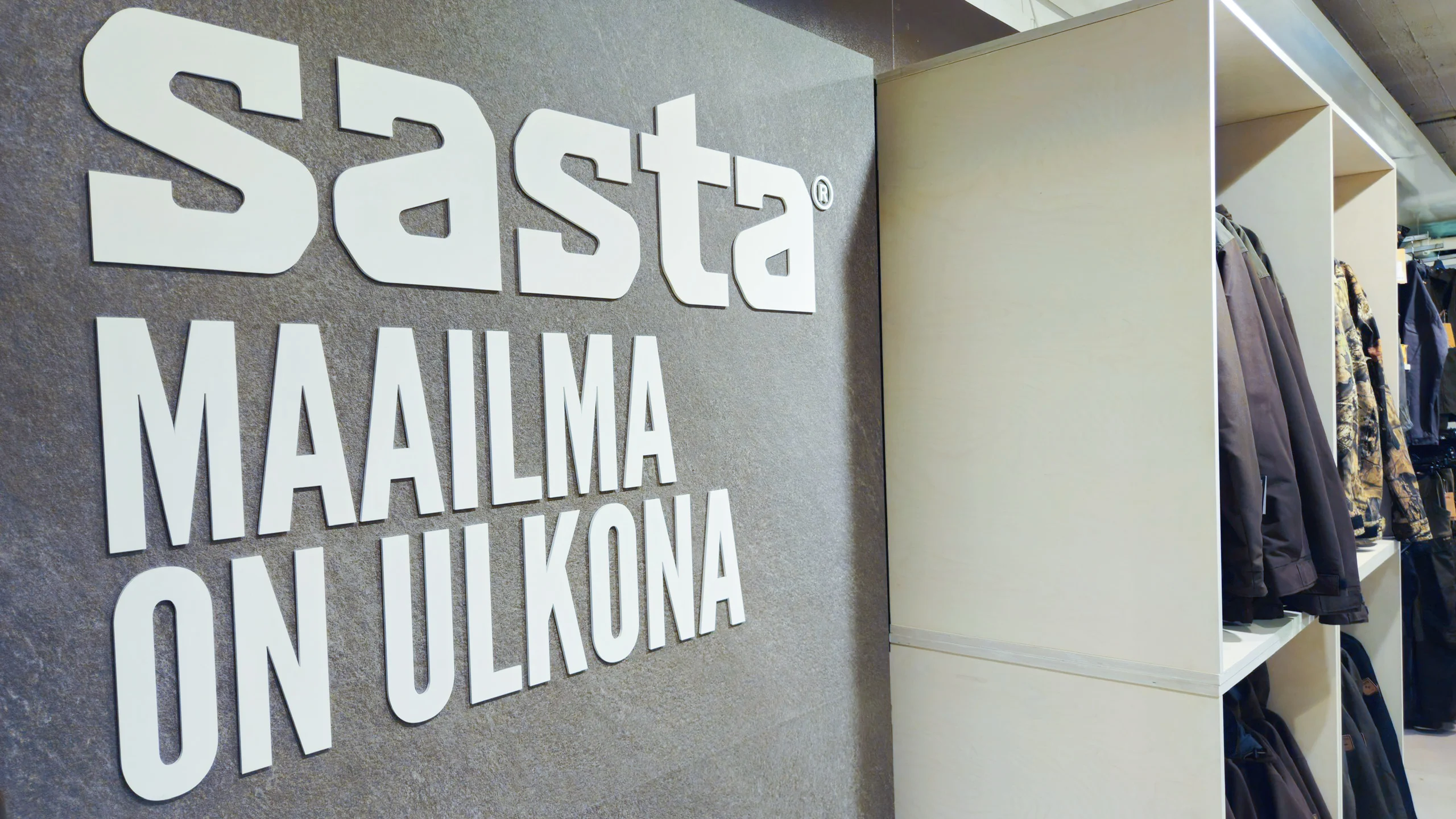 Sasta-logo ja Maailma on ulkona -teksti vaateosaston seinässä