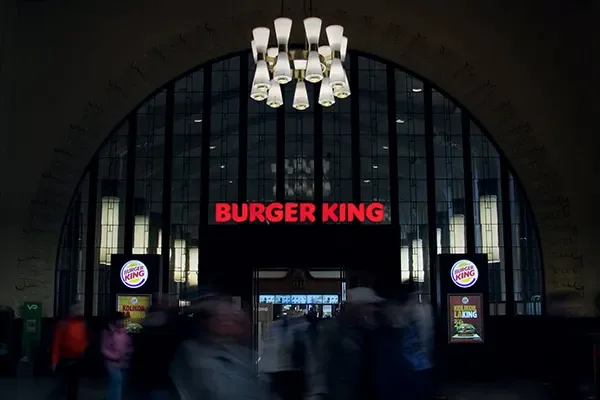 A red illuminated Burger King sign beneath a large arched window in a busy interior.