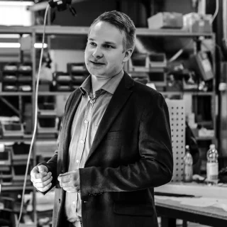 Mikko Okkonen standing in a production workshop in a black-and-white portrait.