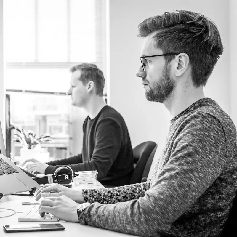 Two people working at computers in an office, shown in black and white.
