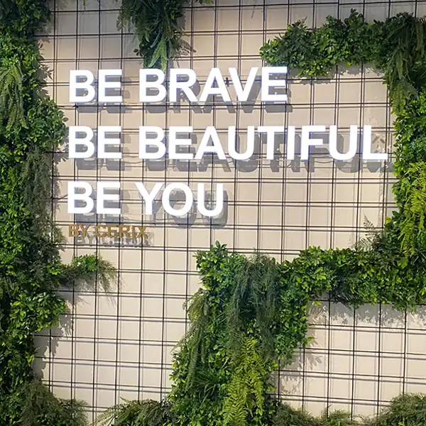 Illuminated words “Be brave, be beautiful, be you” on a wire grid decorated with plants.